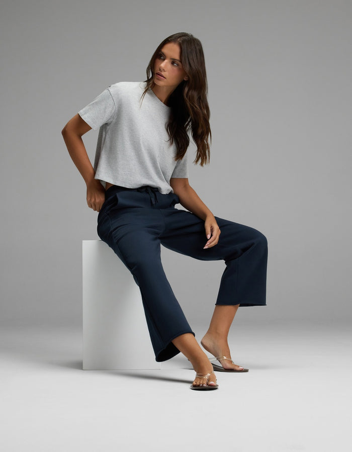 A woman wearing Cozy Earth’s Women’s Chelsea Cropped Pant in navy and a gray t-shirt sits on a white block against a plain gray background, looking to the side with one hand on her hip and sandals on her feet.|Color:Eclipse