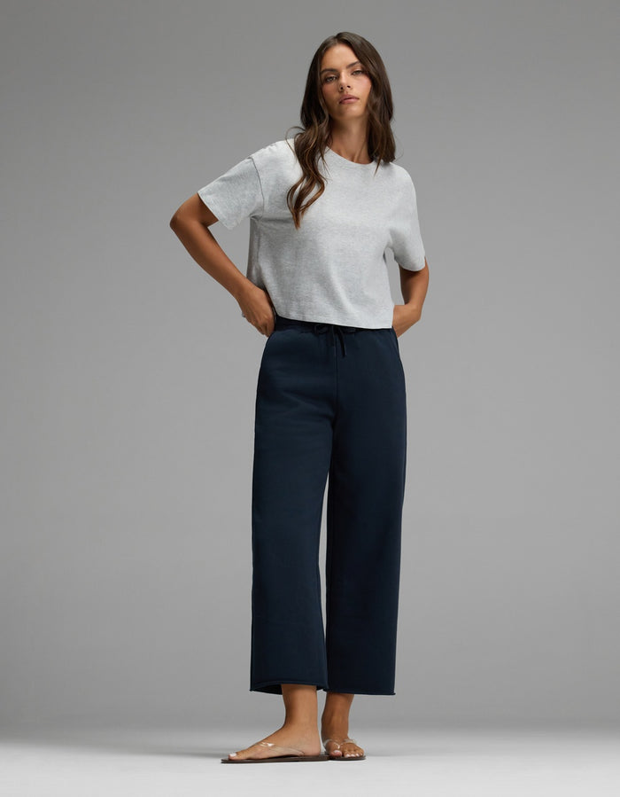 A woman stands barefoot on a gray floor, wearing a light gray short-sleeve t-shirt and Cozy Earth’s Women’s Chelsea Cropped Pant in black, with her hands in her pockets as she looks forward against a plain background.|Color:Eclipse