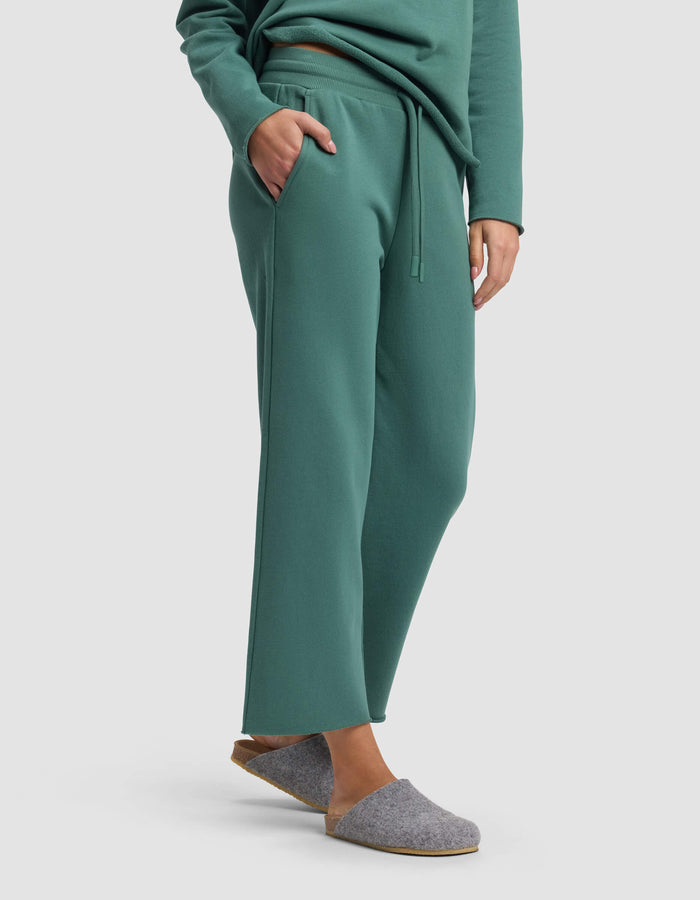 A person stands against a plain background with one hand in their pocket, wearing Cozy Earth's Women's Chelsea Cropped Pant in green, paired with a matching green sweatshirt and gray felt slippers.|Color:Sea Green