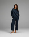 A woman stands in a relaxed pose, hands in her pockets, wearing the Cozy Earth Women’s Chelsea Cropped Set—a matching navy blue long-sleeve top and wide-leg pants—with brown sandals, against a plain gray background.|Color:Eclipse