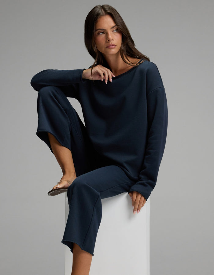 Wearing the Cozy Earth Women’s Chelsea Cropped Set, a woman sits on a white block with one knee raised and the other leg hanging down, gazing to the side against a plain gray background.|Color:Eclipse