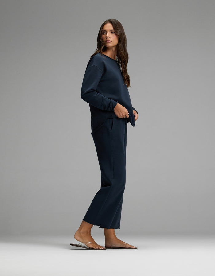 A woman stands sideways against a plain gray background, wearing the Women’s Chelsea Cropped Set in navy blue from Cozy Earth, paired with clear sandals.|Color:Eclipse