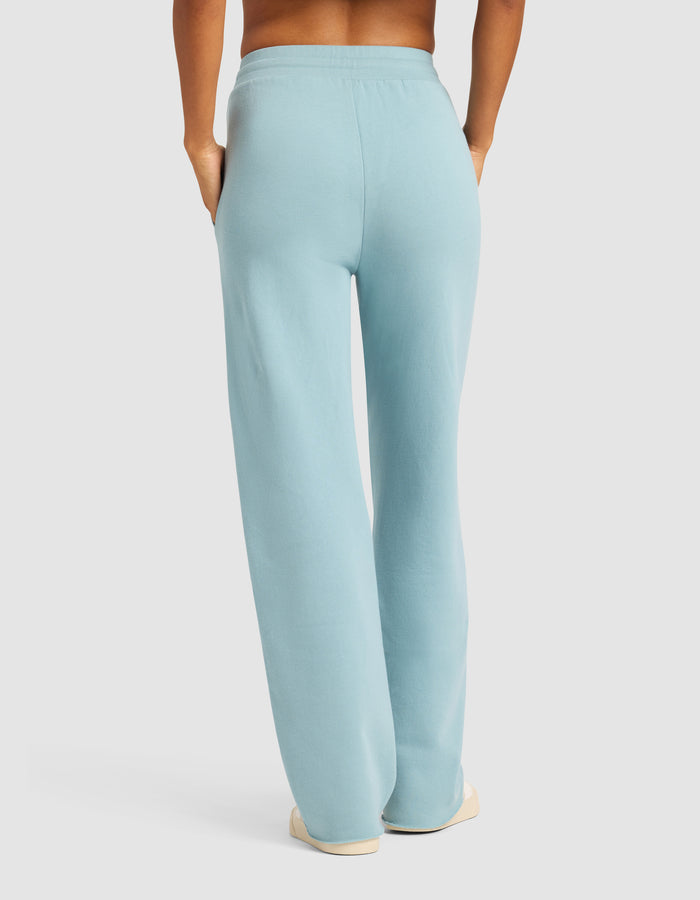Model wearing Cozy Earth Chelsea Pant in light blue, paired with white shoes. Shown from the back with hands in pockets against a plain light background.|Color:River