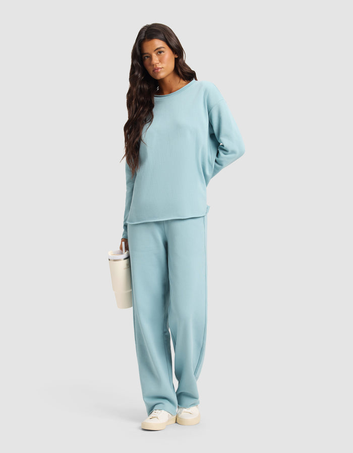 A woman with long dark hair wears the Cozy Earth Chelsea Pant, styled with a light blue long-sleeve top and beige sneakers. She holds a white tumbler and stands against a plain light gray background.|Color:River