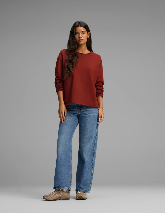 A woman with long dark hair wears the Cozy Earth Women's Chelsea Pullover in a rust shade, paired with blue jeans and brown sneakers. She stands against a plain gray background, looking at the camera with a relaxed posture.|Color:Currant