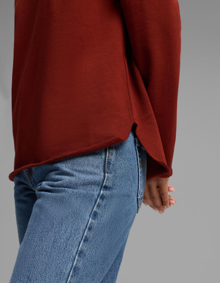 A person shown from the waist down, standing sideways with one hand behind their back against a plain gray background, wearing blue jeans and the Women's Chelsea Pullover in rust red by Cozy Earth.|Color:Currant