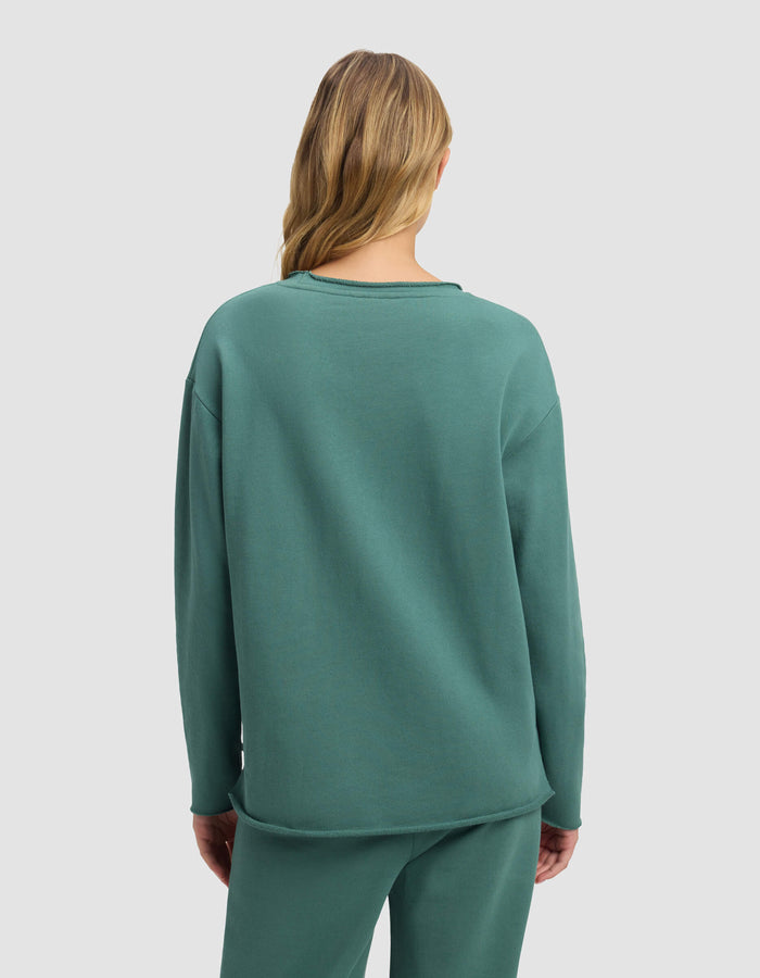 A woman with long, wavy blonde hair is seen from behind wearing the Cozy Earth Women's Chelsea Pullover in teal, paired with matching pants, set against a plain light gray background.|Color:Sea Green