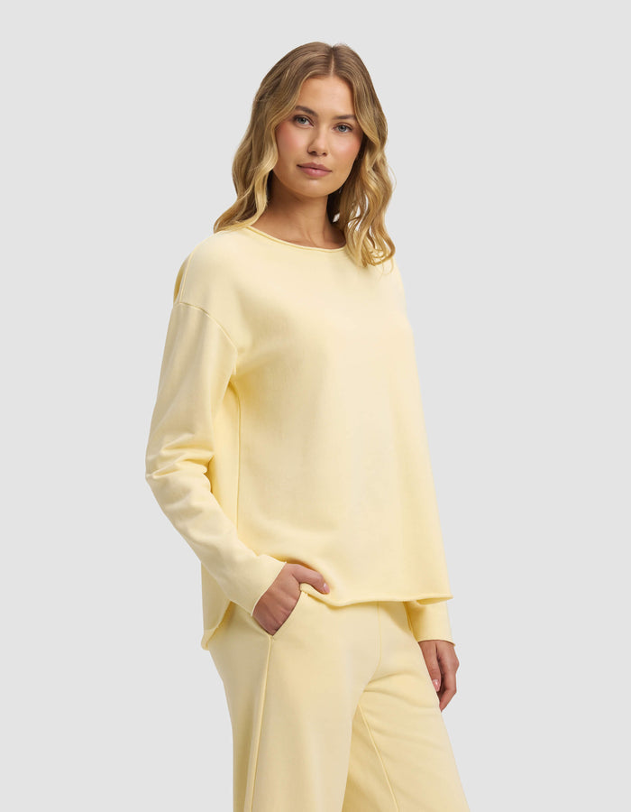 A woman with wavy blonde hair models the Cozy Earth Women's Chelsea Pullover and matching pants in pale yellow, standing against a light gray background with one hand in her pocket.|Color:Meringue
