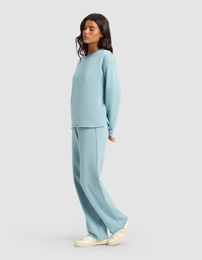 A woman models the Cozy Earth Women's Chelsea Set—loose-fitting, light blue long-sleeve top and matching wide-leg pants—paired with white sneakers, standing sideways against a plain light background.|Color:River