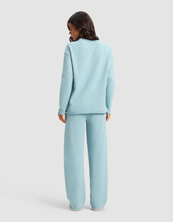 A woman faces away, wearing the Cozy Earth Women's Chelsea Set—loose-fitting light blue sweatshirt and matching wide-leg pants—against a plain light background.|Color:River