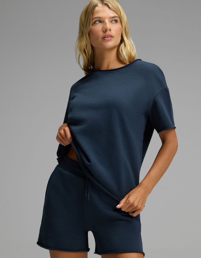A woman stands against a plain background wearing the Cozy Earth Women’s Chelsea Short Sleeve Set in navy blue. She holds the side of her shirt and looks slightly to her left.|Color:Eclipse