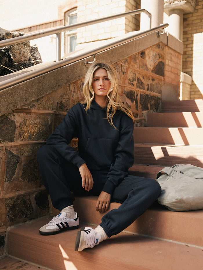 A person with long, blonde hair sits on outdoor steps made of brick and concrete. They are wearing a dark Women's CityScape Cropped Hoodie from Cozy Earth and matching sweatpants, paired with white Adidas sneakers. A grey backpack is placed beside them. The person has a serious expression. |Color:Eclipse