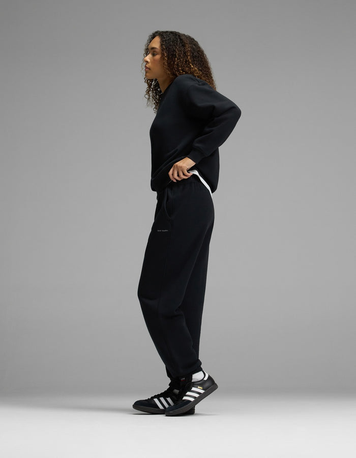 A person with curly hair stands sideways in a relaxed pose, wearing the Women’s CityScape Crewneck & Sweatpant Set by Cozy Earth in black, paired with black Adidas sneakers featuring white stripes, against a plain gray background.|Color:Jet Black