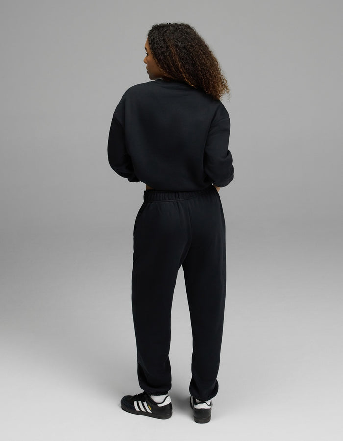 A person with curly hair, seen from the back, is wearing the Women’s CityScape Crewneck & Sweatpant Set by Cozy Earth in black, along with black Adidas sneakers featuring white stripes, standing on a light gray background.|Color:Jet Black