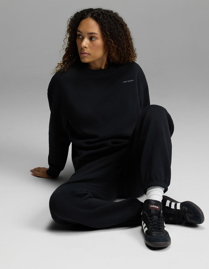 A woman with curly hair sits on the floor with one leg bent, looking to the side against a plain gray background, wearing the Women’s CityScape Crewneck & Sweatpant Set from Cozy Earth.|Color:Jet Black