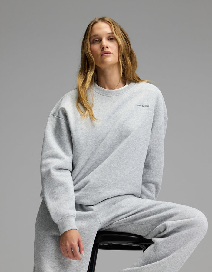 A woman with long blonde hair sits on a stool, wearing the Cozy Earth Women’s CityScape Crewneck & Sweatpant Set in light gray, against a plain gray background.|Color:Heather Grey