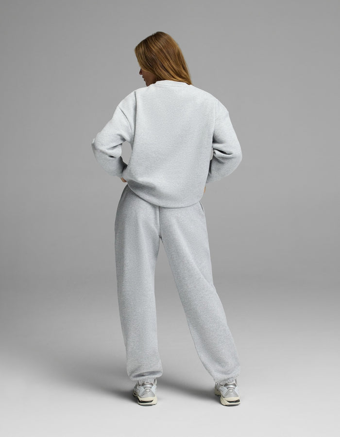 A person stands facing away, wearing the Cozy Earth Women’s CityScape Crewneck & Sweatpant Set in light gray. They have long hair, white sneakers, and their hands rest on their hips against a plain gray background.|Color:Heather Grey