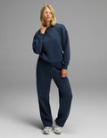 A woman with shoulder-length blonde hair stands against a gray background wearing the Cozy Earth Women’s Cityscape Crewneck & Wide Leg Pant Set in Eclipse, paired with silver sneakers. She holds her left hand near her chin.|Color:Eclipse