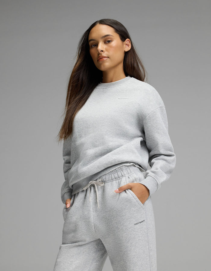 A woman with long brown hair models the Cozy Earth Women’s Cityscape Crewneck & Wide Leg Pant Set in light gray, standing with one hand in her pocket against a plain gray background.|Color:Heather Grey