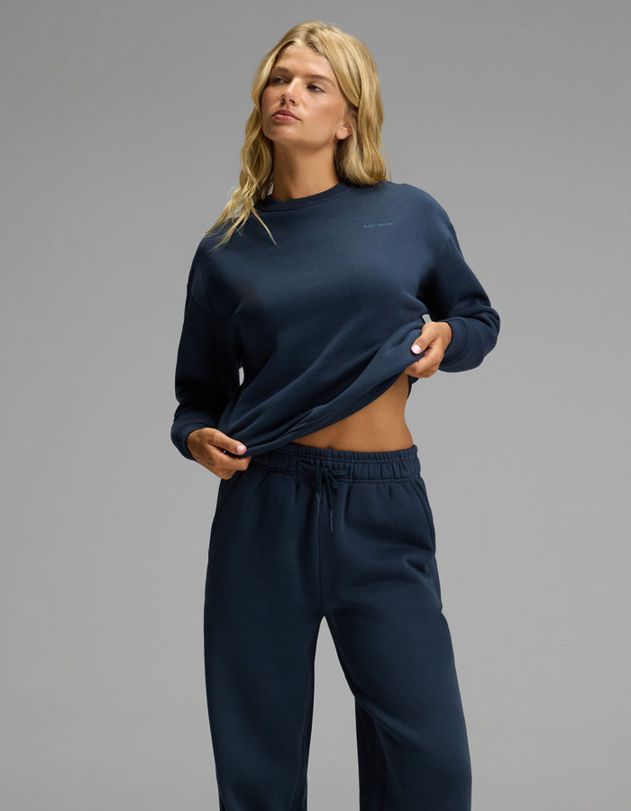 A person with long blonde hair wears the Cozy Earth Women’s Cityscape Crewneck & Wide Leg Pant Set in dark blue, holding up the hem of the crewneck slightly to reveal their midriff against a plain gray background.|Color:Eclipse