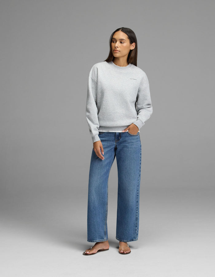 A woman stands against a gray background wearing the Cozy Earth Women’s Cityscape Crewneck, styled with blue wide-leg jeans and beige sandals. Her left hand is in her pocket as she looks slightly to the side.|Color:Heather Grey