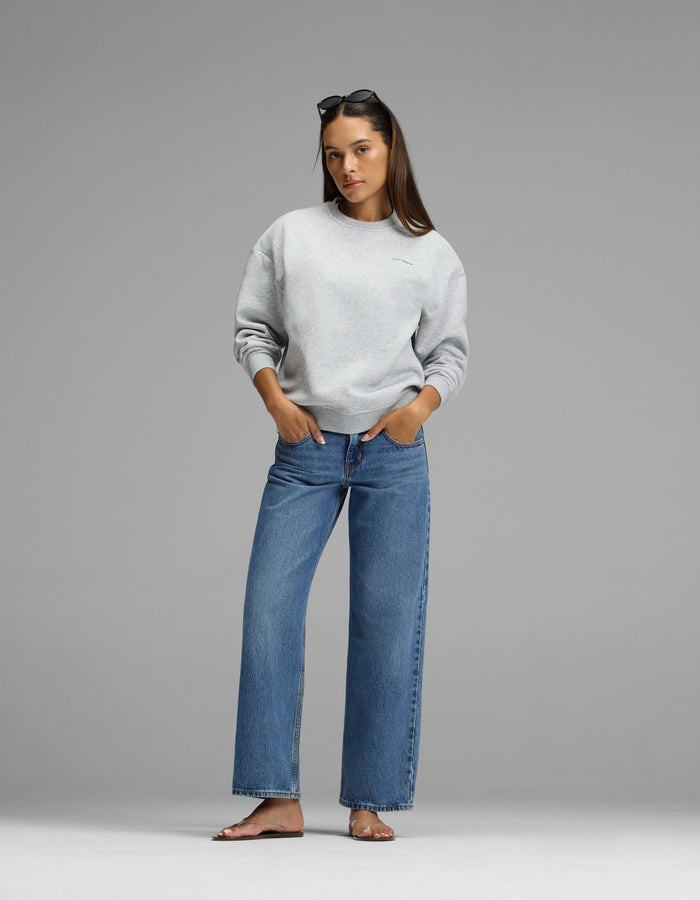 A woman stands against a gray background, wearing the Cozy Earth Women’s Cityscape Crewneck in light gray, paired with blue jeans and sunglasses on her head. Her hands are in her pockets and she is barefoot.|Color:Heather Grey