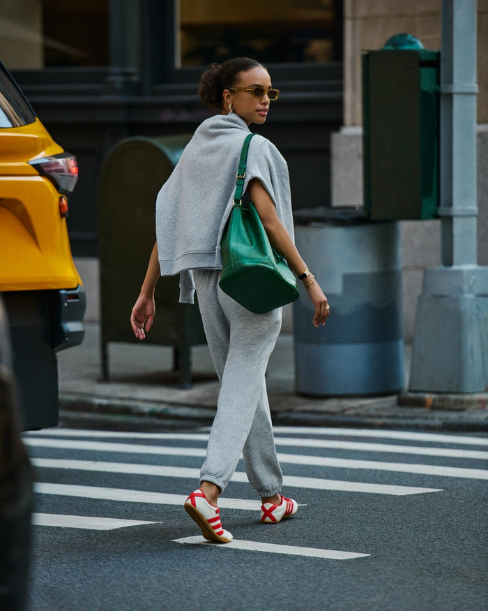 A woman walking across a city crosswalk wearing a light gray Cozy Earth Cityscape Crewneck around her shoulders and Cozy Earth Cityscape sweatpants, a green shoulder bag, and red-and-white sneakers. A yellow taxi is partially visible on the left, with urban buildings and street elements in the background.|Color:Heather Grey