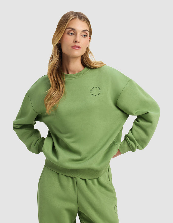 A woman with long blonde hair stands with her hands on her hips against a plain light gray background, wearing the Cozy Earth Women's Cityscape Crewneck in a loose-fitting, light green style with matching sweatpants.|Color:Clover
