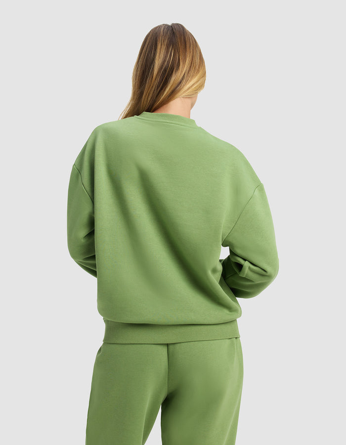 A woman with long, light brown hair is seen from behind wearing the Cozy Earth Women's Cityscape Crewneck in light green, paired with matching sweatpants, standing against a plain light gray background.|Color:Clover