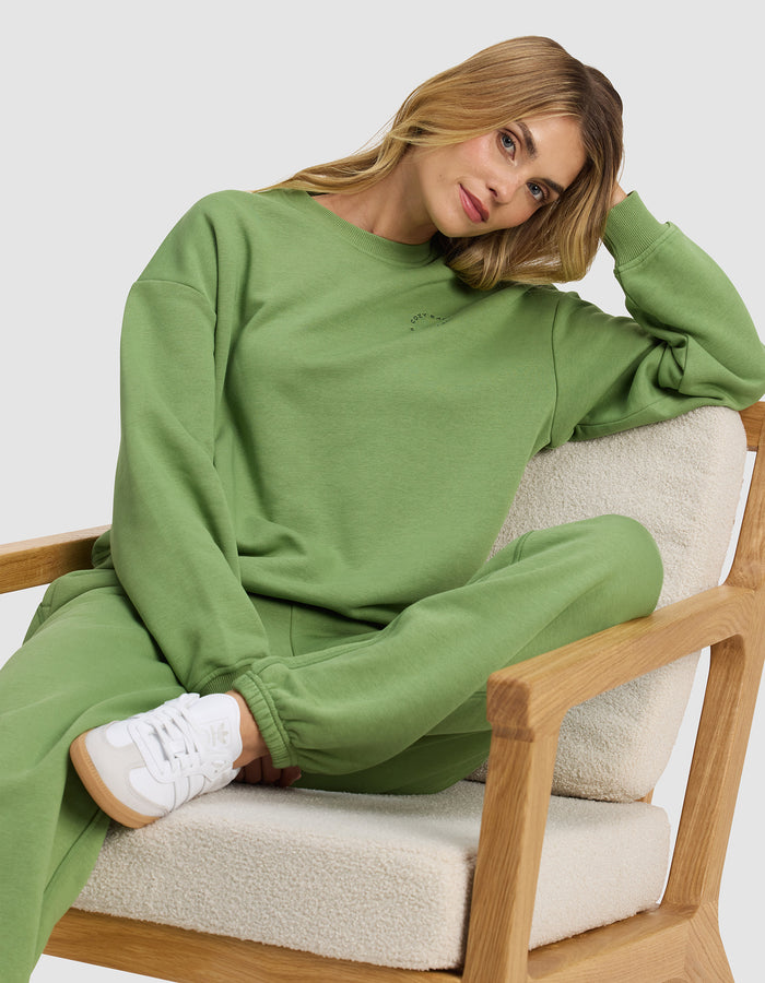 A woman wears the Cozy Earth Women's Cityscape Crewneck, lounging casually on a cushioned wooden chair and looking relaxed in her stylish, comfortable outfit.|Color:Clover