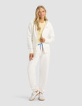 A woman models the Cozy Earth Women's CityScape Full Zip & Sweatpant Set in white, paired with a light yellow top and white sneakers, standing against a plain light gray background.|Color:Sea Salt