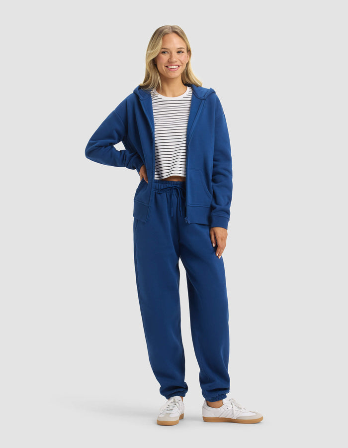 A woman smiles in Cozy Earth's Women's CityScape Full Zip & Sweatpant Set—blue zip-up hoodie and matching sweatpants—paired with a white and navy striped tee and white sneakers, set against a light gray background.|Color:Pacific