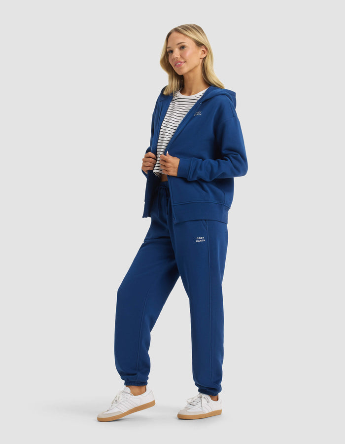 A woman wears the Cozy Earth Women's CityScape Full Zip & Sweatpant Set, paired with white sneakers and a striped shirt, smiling and looking to the side against a plain light background.|Color:Pacific