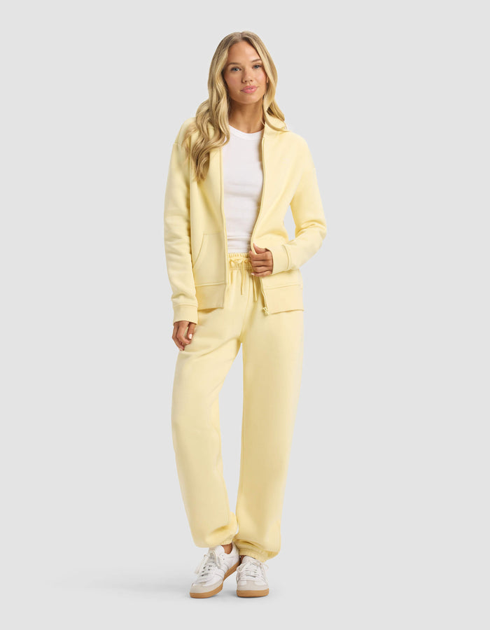 A woman with long blonde hair wears the Cozy Earth Women's CityScape Full Zip & Sweatpant Set in light yellow, paired with a white t-shirt and sneakers. She stands facing forward against a plain light gray background.|Color:Meringue