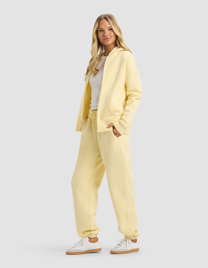 A woman with long blonde hair smiles in Cozy Earth’s Women's CityScape Full Zip & Sweatpant Set, paired with white sneakers and a white top, standing against a plain light gray background.|Color:Meringue