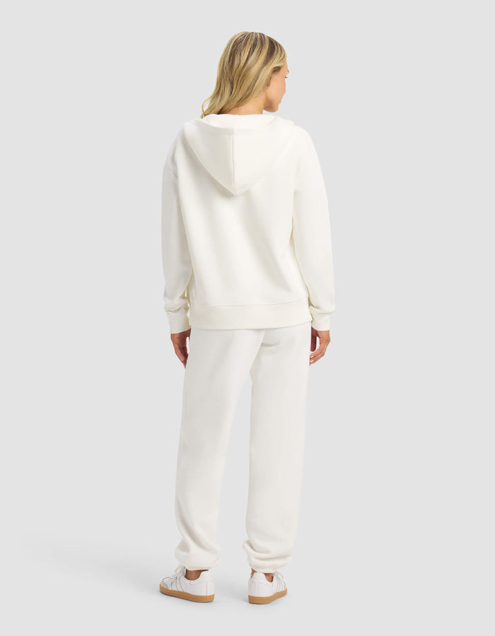 A woman with blonde hair faces away, wearing the Cozy Earth Women's CityScape Full Zip & Sweatpant Set in white with matching sneakers, against a plain light gray background.|Color:Sea Salt