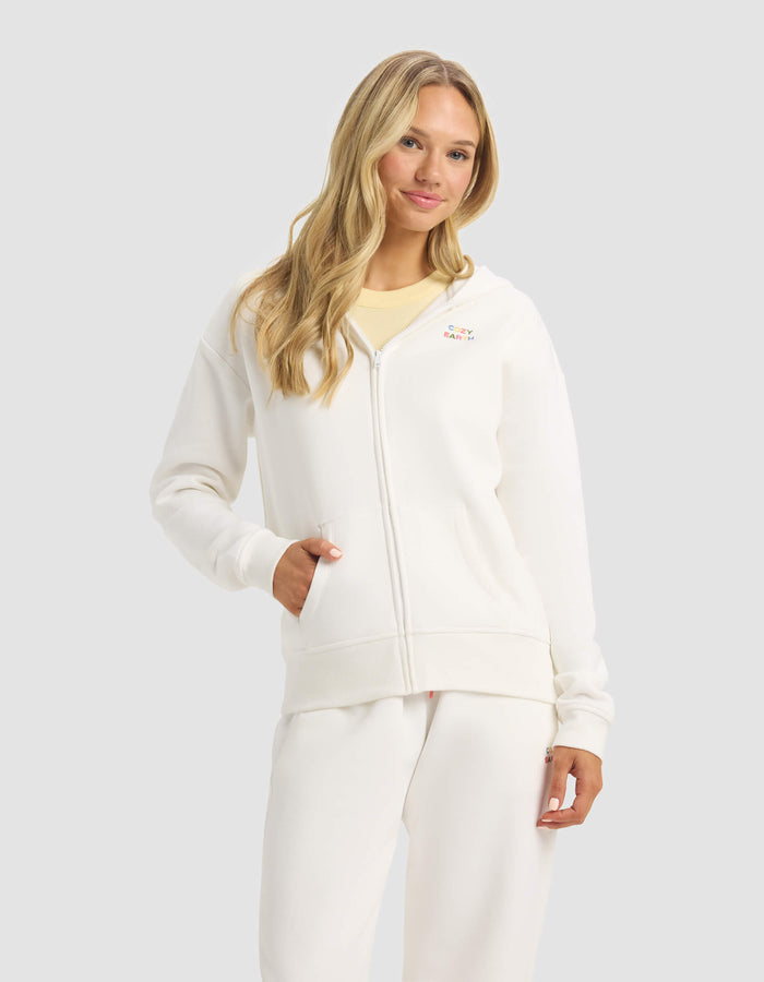 A woman with long blonde hair models the Cozy Earth Women's CityScape Full Zip hoodie and matching white sweatpants against a light gray background, one hand in her hoodie pocket.|Color:Sea Salt