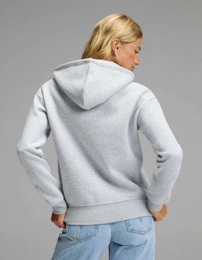 A woman with blonde hair, wearing the Cozy Earth Women’s CityScape Full Zip Hoodie in light gray and blue jeans, stands facing away from the camera against a plain gray background.