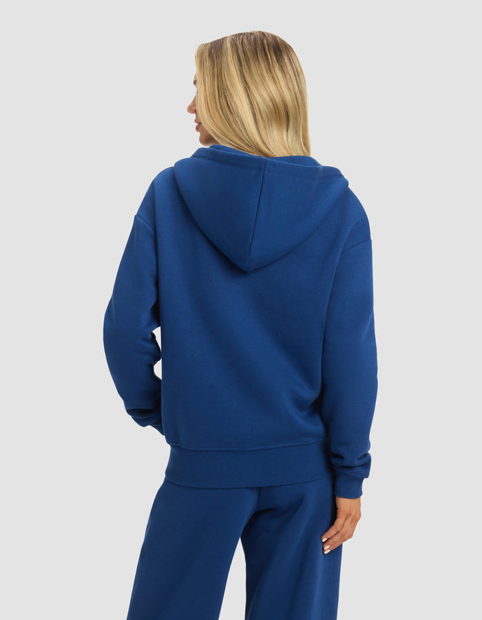 A person with long blonde hair is shown from the back, wearing the Cozy Earth Women's CityScape Full Zip in blue with matching pants against a plain light gray background.|Color:Pacific