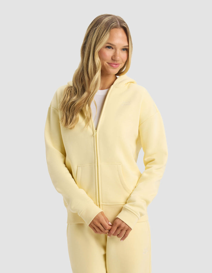 A woman with long blonde hair wears the Cozy Earth Women's CityScape Full Zip hoodie and matching sweatpants, standing with her hands together and smiling slightly against a plain light gray background.|Color:Meringue