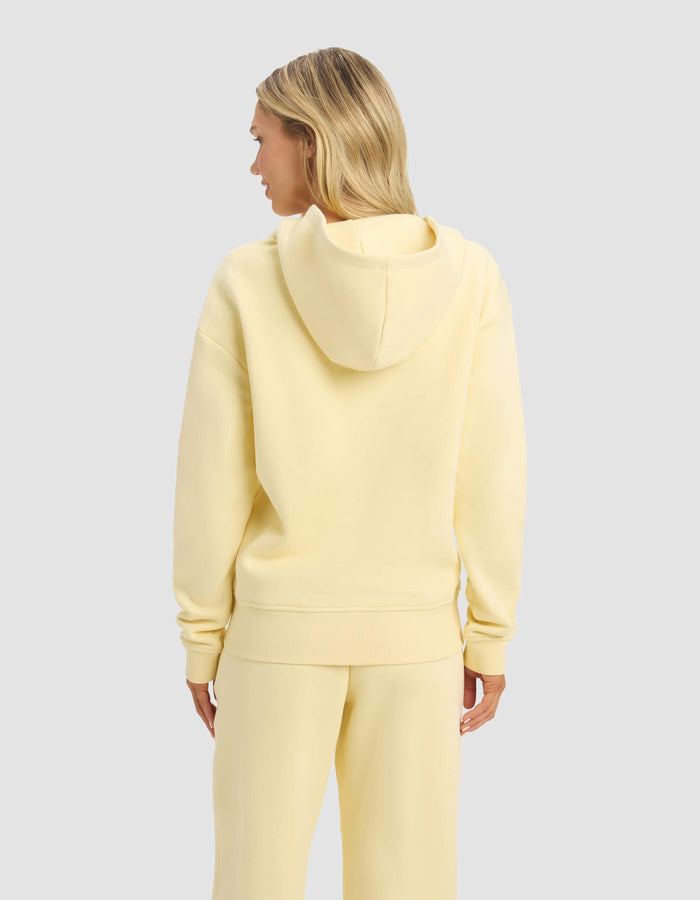 A person with long blonde hair is seen from behind wearing the Cozy Earth Women's CityScape Full Zip hoodie and matching pants in pale yellow, standing against a plain light gray background.|Color:Meringue