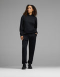 A person with curly hair stands against a gray background, wearing the Cozy Earth Women’s CityScape Hoodie & Sweatpant Set in black, paired with black sneakers and one hand in their pocket.|Color:Jet Black