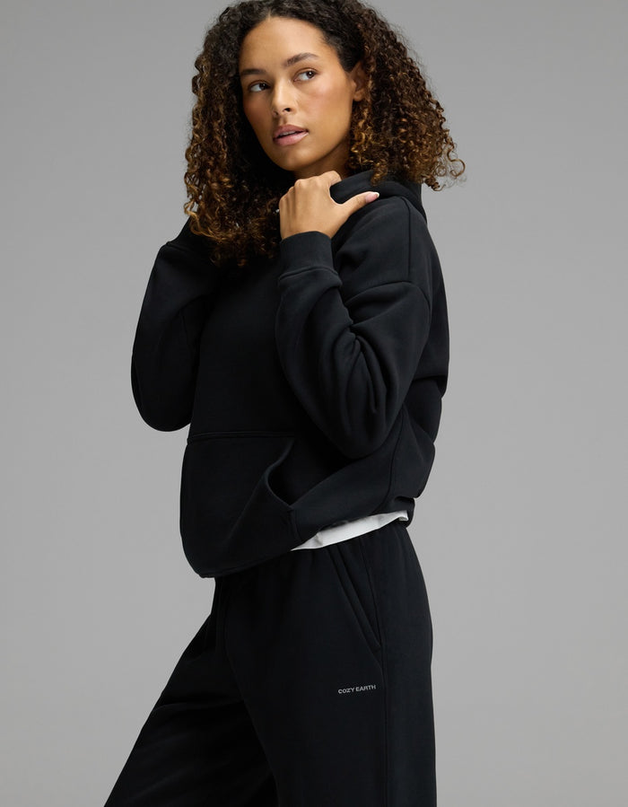 A woman with curly hair wears the Cozy Earth Women’s CityScape Hoodie & Sweatpant Set in black, standing against a neutral gray background and looking to the side as she adjusts her hood.|Color:Jet Black