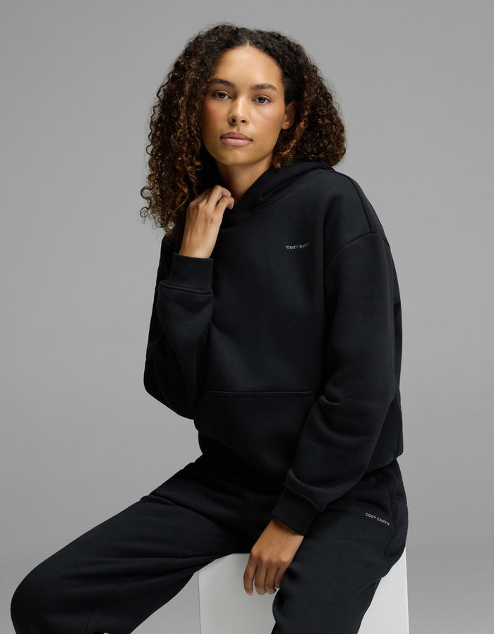A woman with curly hair models the Cozy Earth Women’s CityScape Hoodie & Sweatpant Set in black, sitting on a white cube against a plain gray background and looking calmly at the camera.|Color:Jet Black