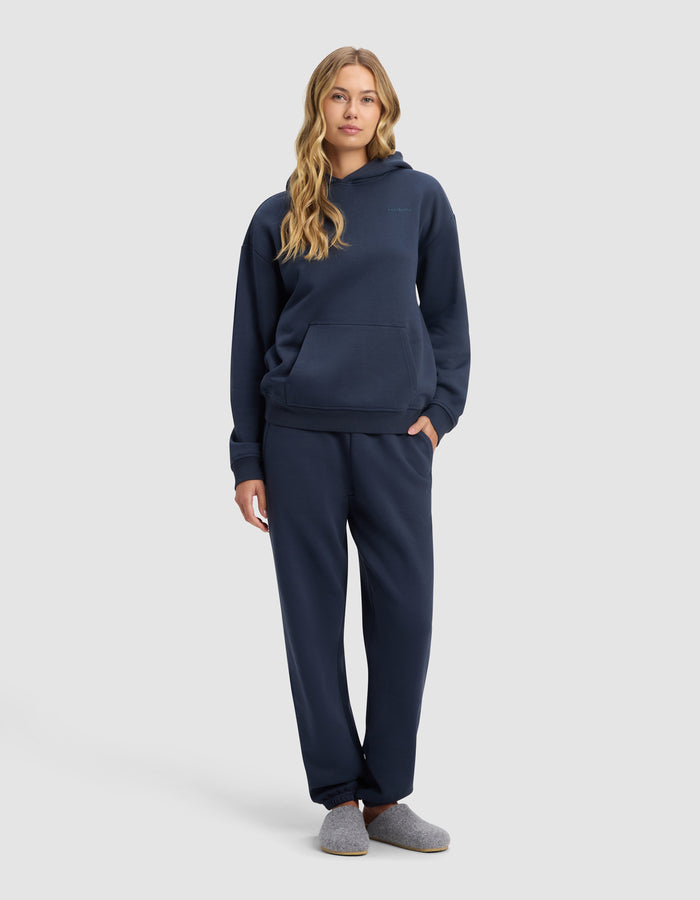 A woman with long wavy hair stands facing forward, wearing Cozy Earth’s Women’s Cityscape Hoodie & Sweatpant Set in navy blue, paired with gray slippers, against a plain light gray background.|Color:Eclipse