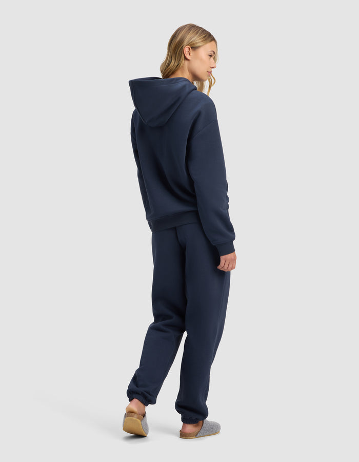 A person with long blonde hair faces away, wearing the Cozy Earth Women's Cityscape Hoodie & Sweatpant Set in navy blue, styled with gray slippers against a plain light gray background.|Color:Eclipse