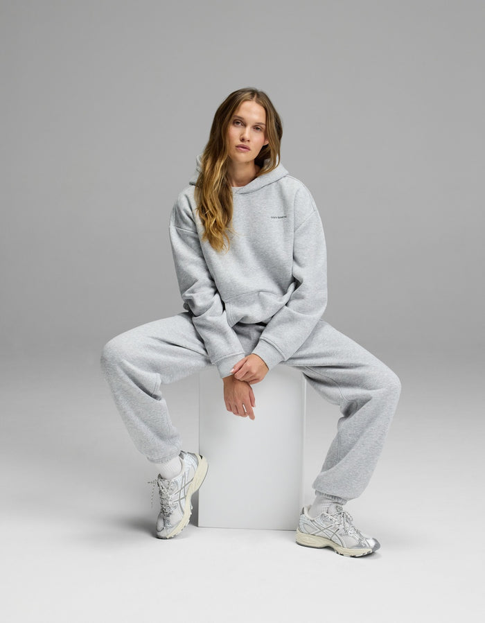Wearing the Cozy Earth Women’s CityScape Hoodie & Sweatpant Set, a woman sits on a white cube against a plain gray background in white sneakers, gazing at the camera with a relaxed expression.|Color:Heather Grey