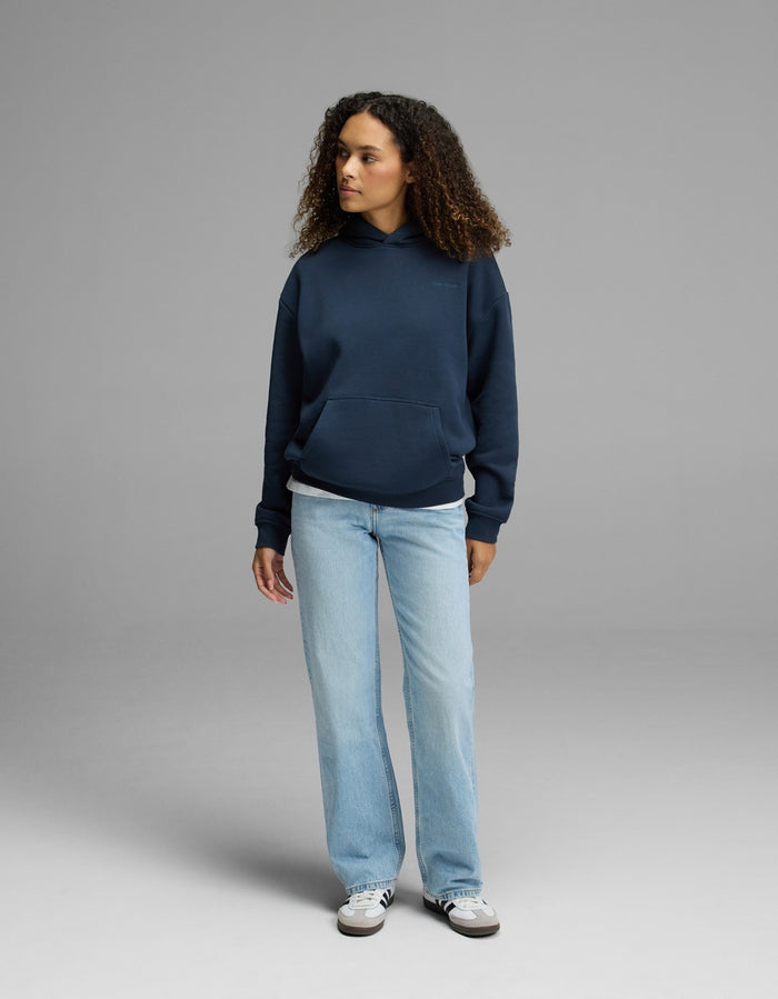 A person with curly hair is wearing the Cozy Earth Women’s CityScape Hoodie in navy blue, paired with light blue jeans and white sneakers, standing against a plain gray background and looking to the side with a neutral expression.|Color:Eclipse