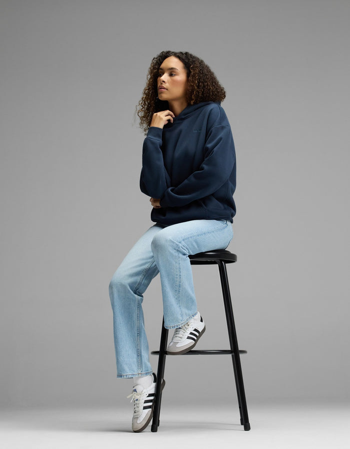 A person with curly hair sits on a tall black stool, wearing a Cozy Earth Women’s CityScape Hoodie in navy, light blue jeans, and white Adidas sneakers, against a plain gray background.|Color:Eclipse