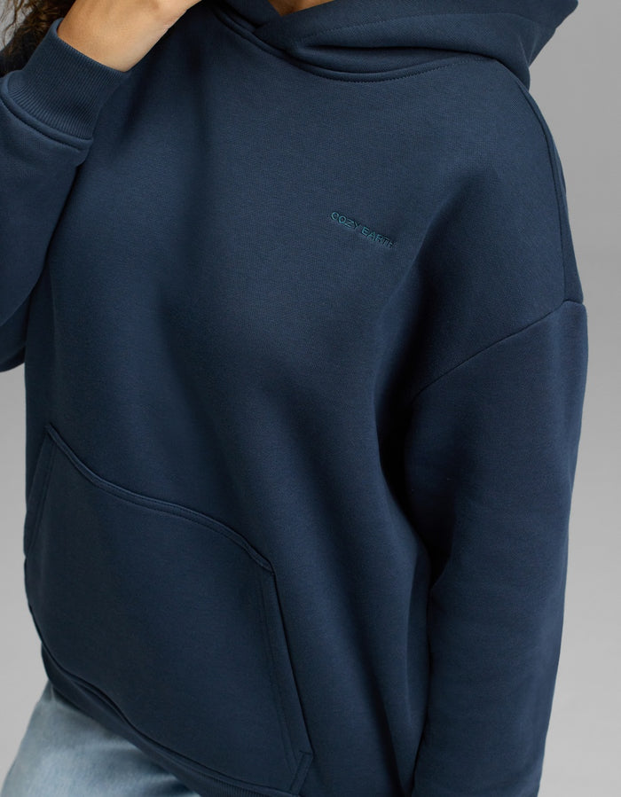 A person is wearing the Cozy Earth Women’s CityScape Hoodie in navy blue, featuring a front pocket and subtle embroidered text on the chest. Their hand rests near their neck against a plain gray background.|Color:Eclipse